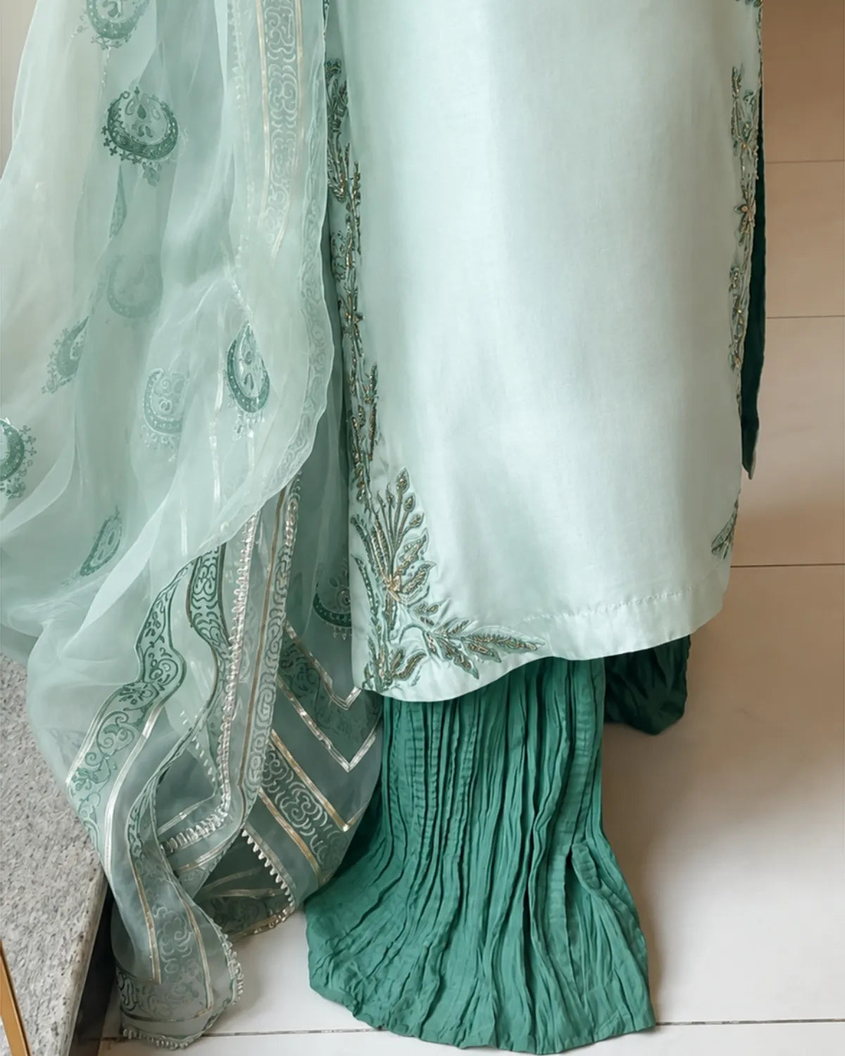 A close-up of a traditional South Asian outfit featuring a mint green embroidered kurta, deep teal pleated palazzo pants, and a matching sheer mint green organza dupatta with gold zari embroidery.