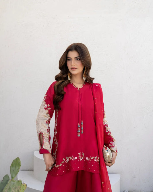 A woman models a bright magenta-pink traditional South Asian outfit with beige floral embroidery on the sleeves and hem, paired with a matching embroidered dupatta and gold jewelry, set against a white textured wall.