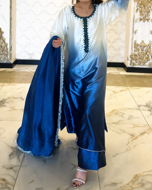 A woman models a traditional three-piece outfit featuring a white and blue gradient satin kurta, a matching blue dupatta with silver embroidery, and silver embellished pants, set against an ornate interior background.