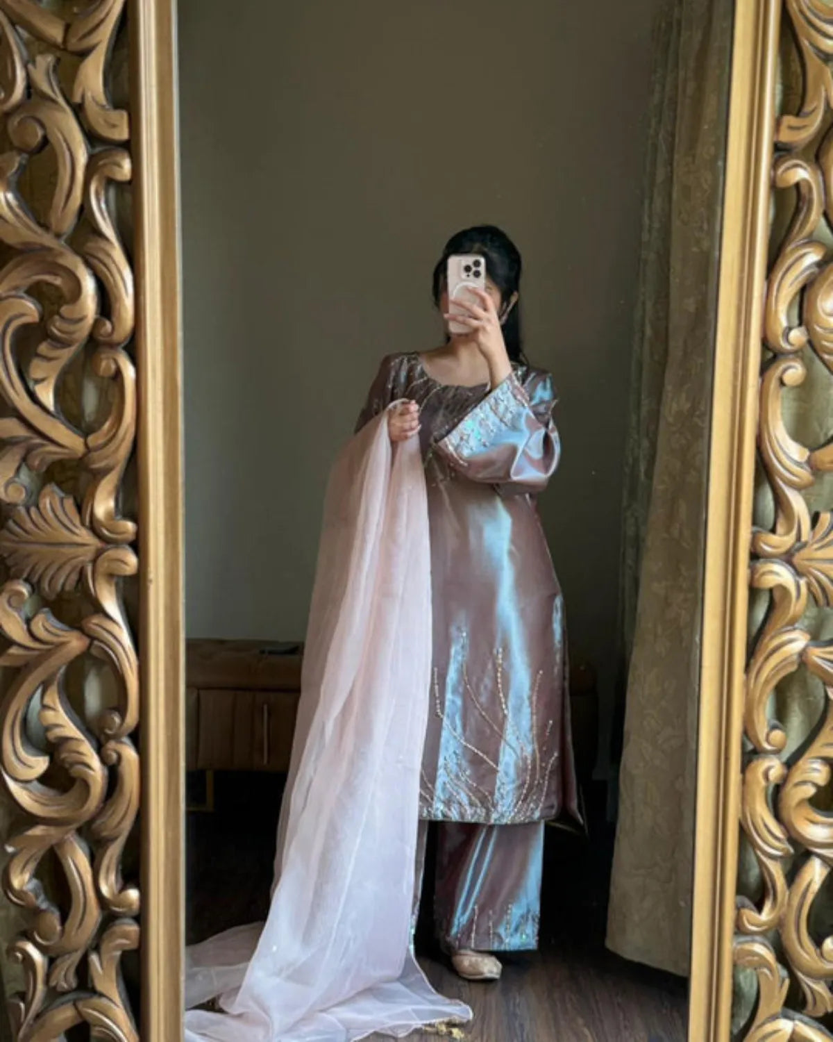 A woman takes a mirror selfie wearing a shimmering metallic lavender-pink traditional outfit with a matching sheer dupatta, framed by an ornate gold mirror.
