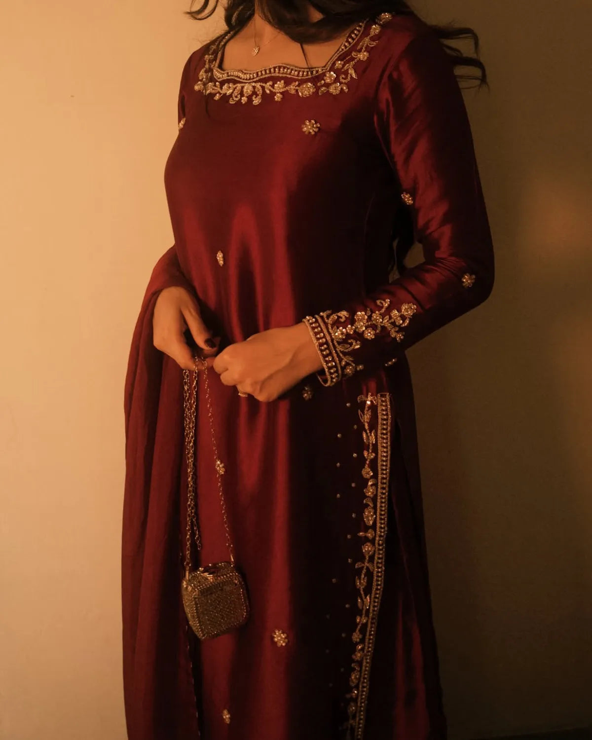A woman wearing a long-sleeved maroon satin dress with gold floral embroidery on the neckline, cuffs, and side borders, holding a small metallic clutch bag against a plain wall.