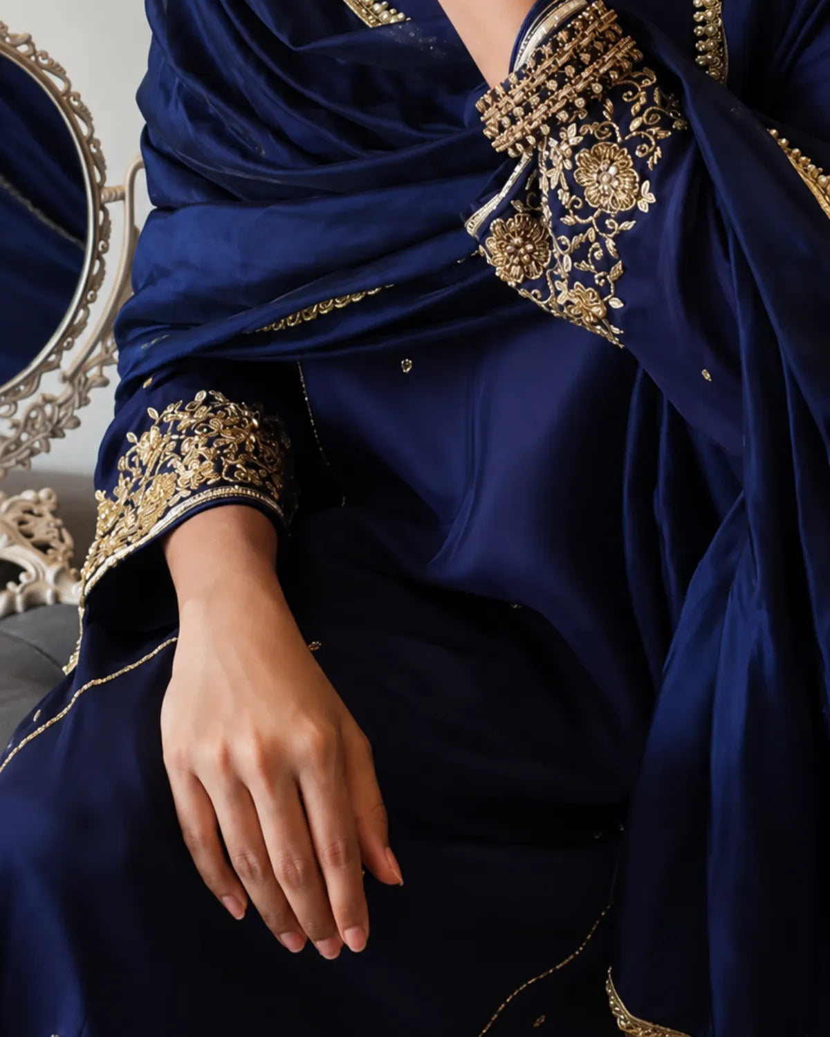 A close-up of a navy blue traditional outfit with ornate gold embroidery on the sleeve cuffs and neckline, worn by a model whose hand is visible resting on the lapel.