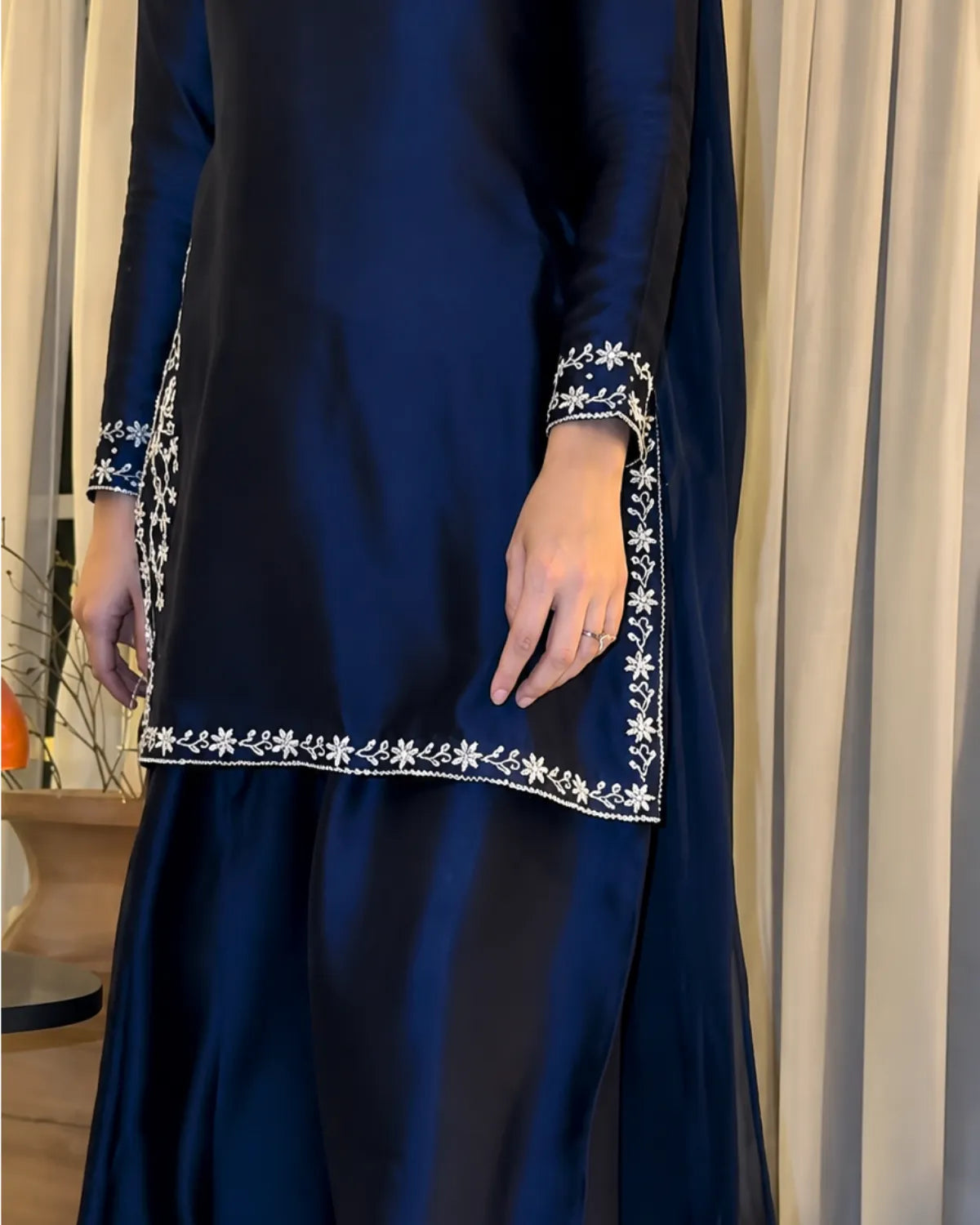 A navy blue traditional outfit with white floral embroidery along the hem and cuffs, worn by a model standing in front of beige curtains.