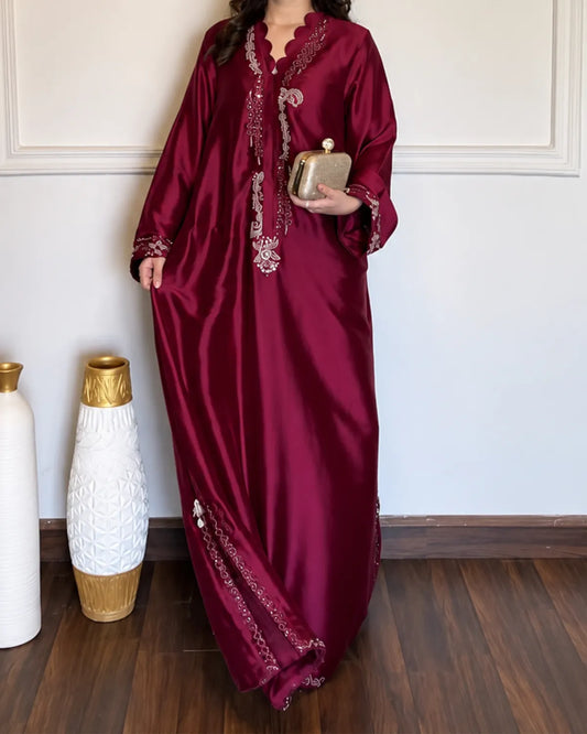A woman wearing a long, flowing maroon satin caftan with intricate silver embroidery, holding a small metallic clutch purse. She is standing in an indoor setting with a white wall and wooden floor, next to two decorative vases.