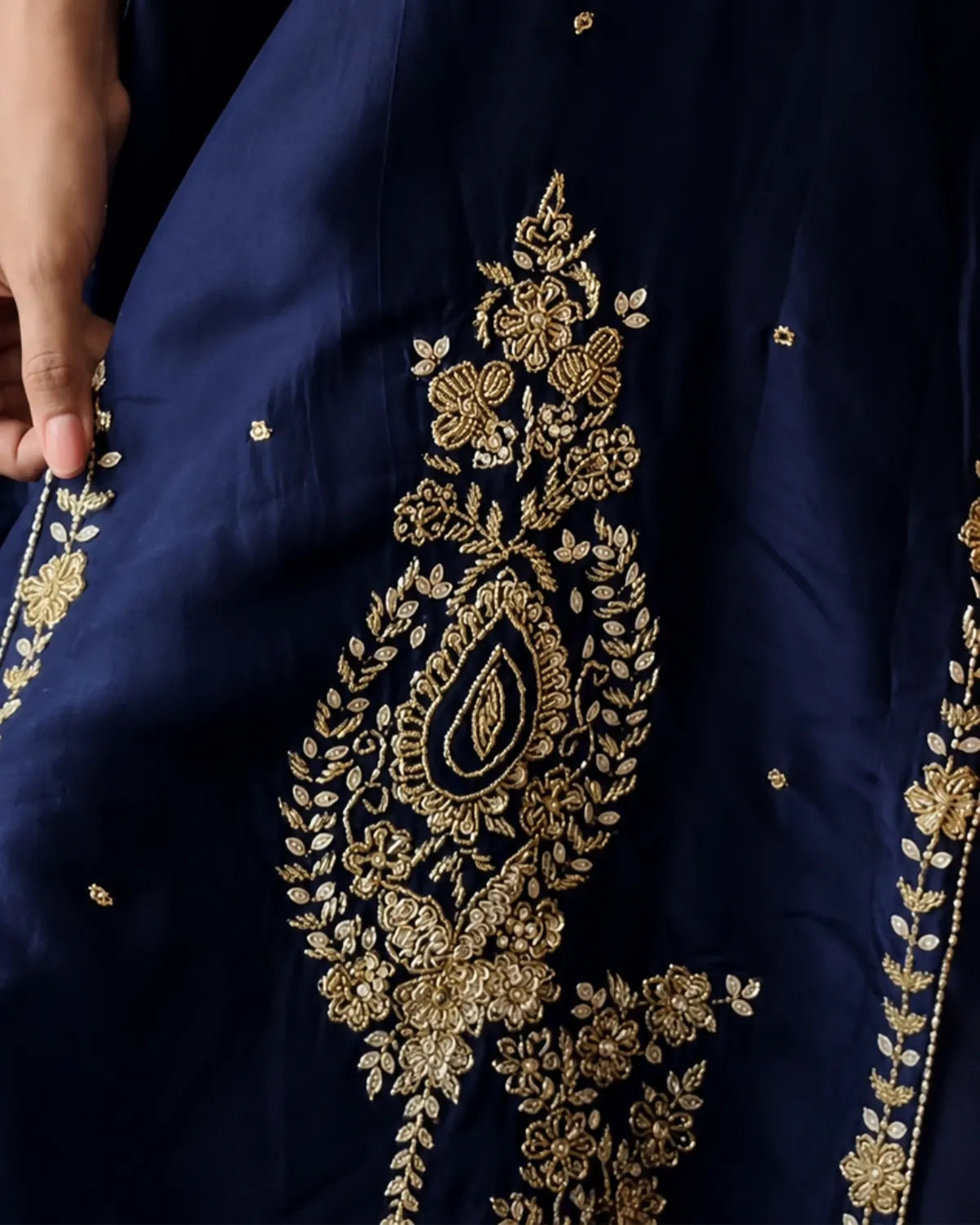 Close-up of a navy blue fabric garment with ornate gold floral and paisley embroidery, showing a hand holding the edge of the fabric.