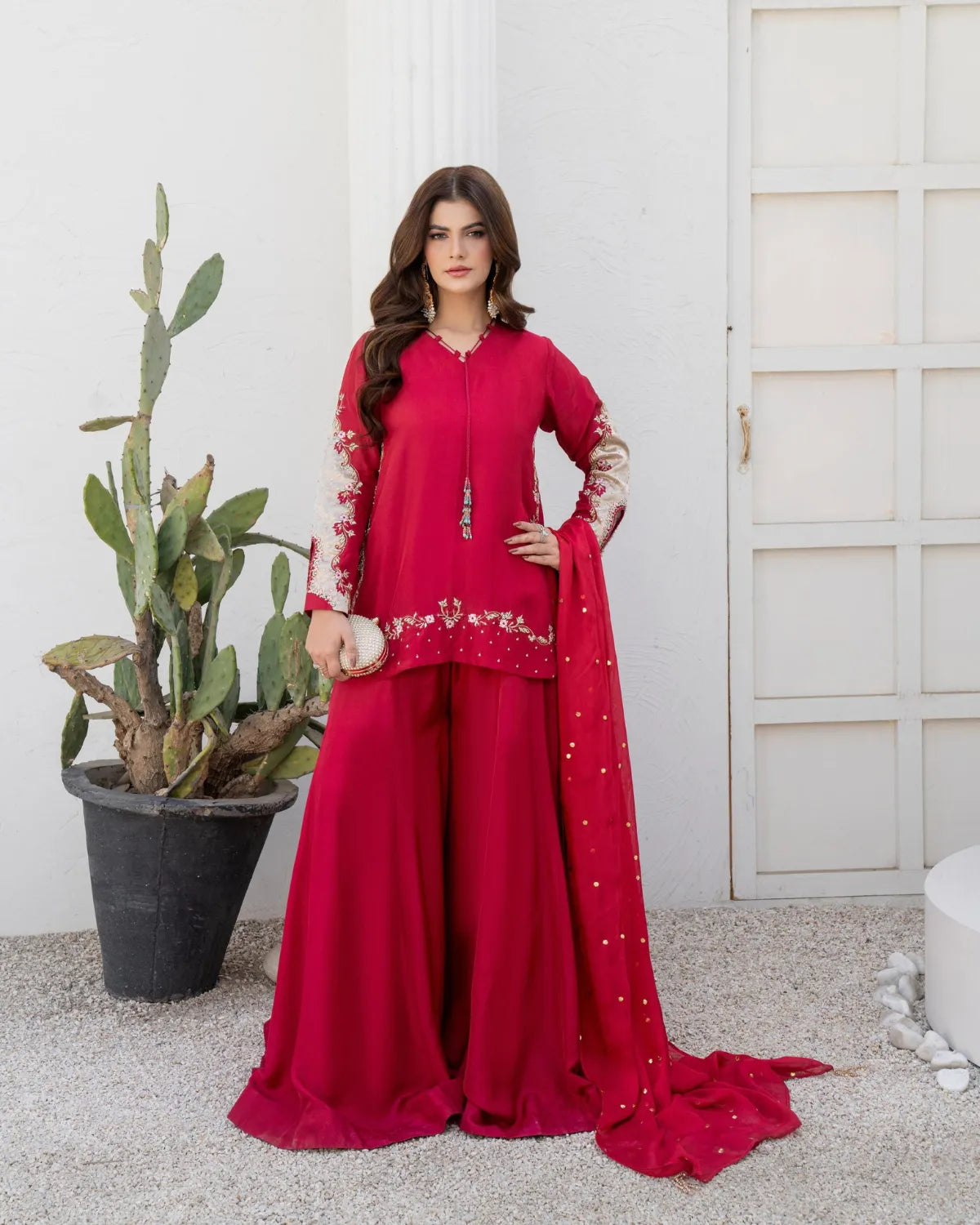 A woman models a vibrant red three-piece traditional outfit with beige floral embroidery on the sleeves and hem, paired with a matching red dupatta, set against a white wall with a potted cactus.