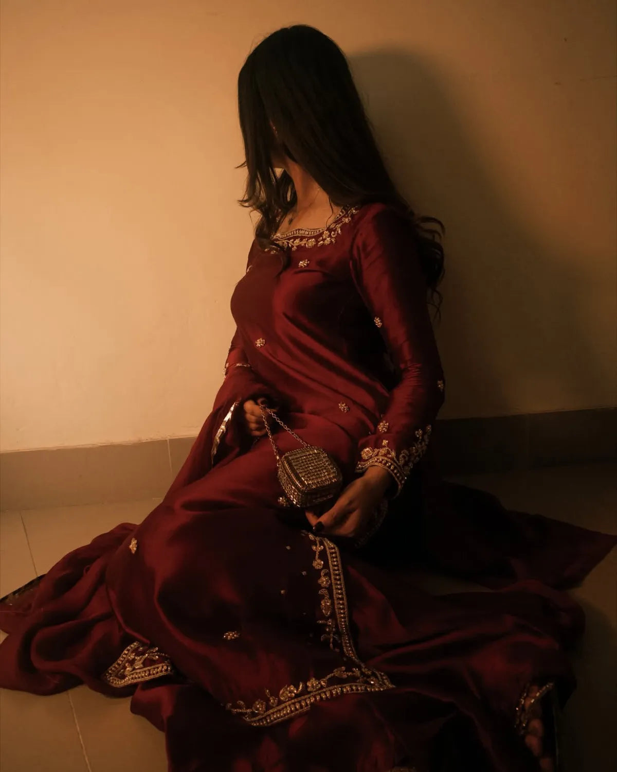 A woman in a deep maroon satin traditional dress with gold embroidery sits on a tiled floor against a plain wall, holding a small metallic clutch bag.