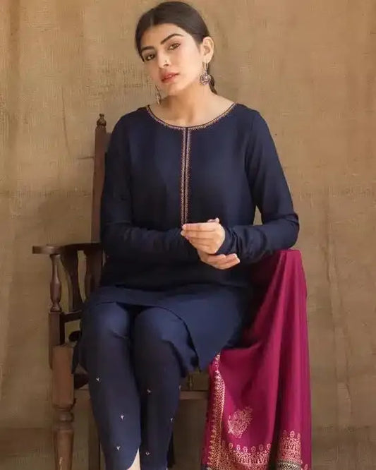 Woman in navy blue traditional outfit with pink dupatta sitting on a wooden chair against a beige wall.