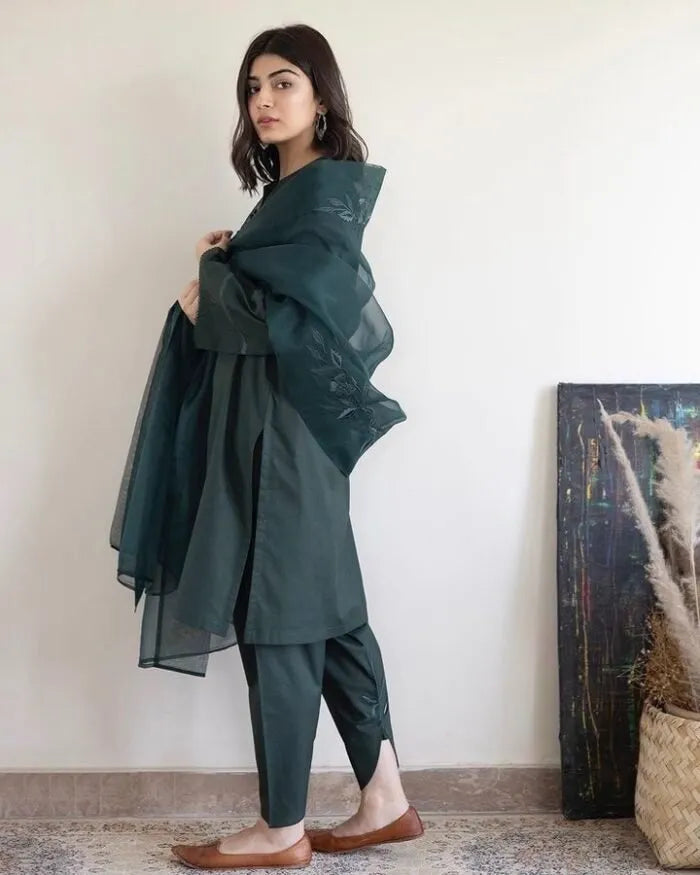 Woman wearing a dark green traditional outfit with a sheer dupatta in a minimal indoor setting.