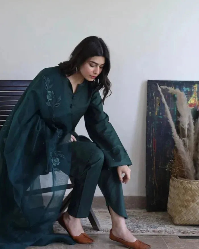 Woman in a dark green outfit sitting on a chair in a room with a painting and plant in the background.