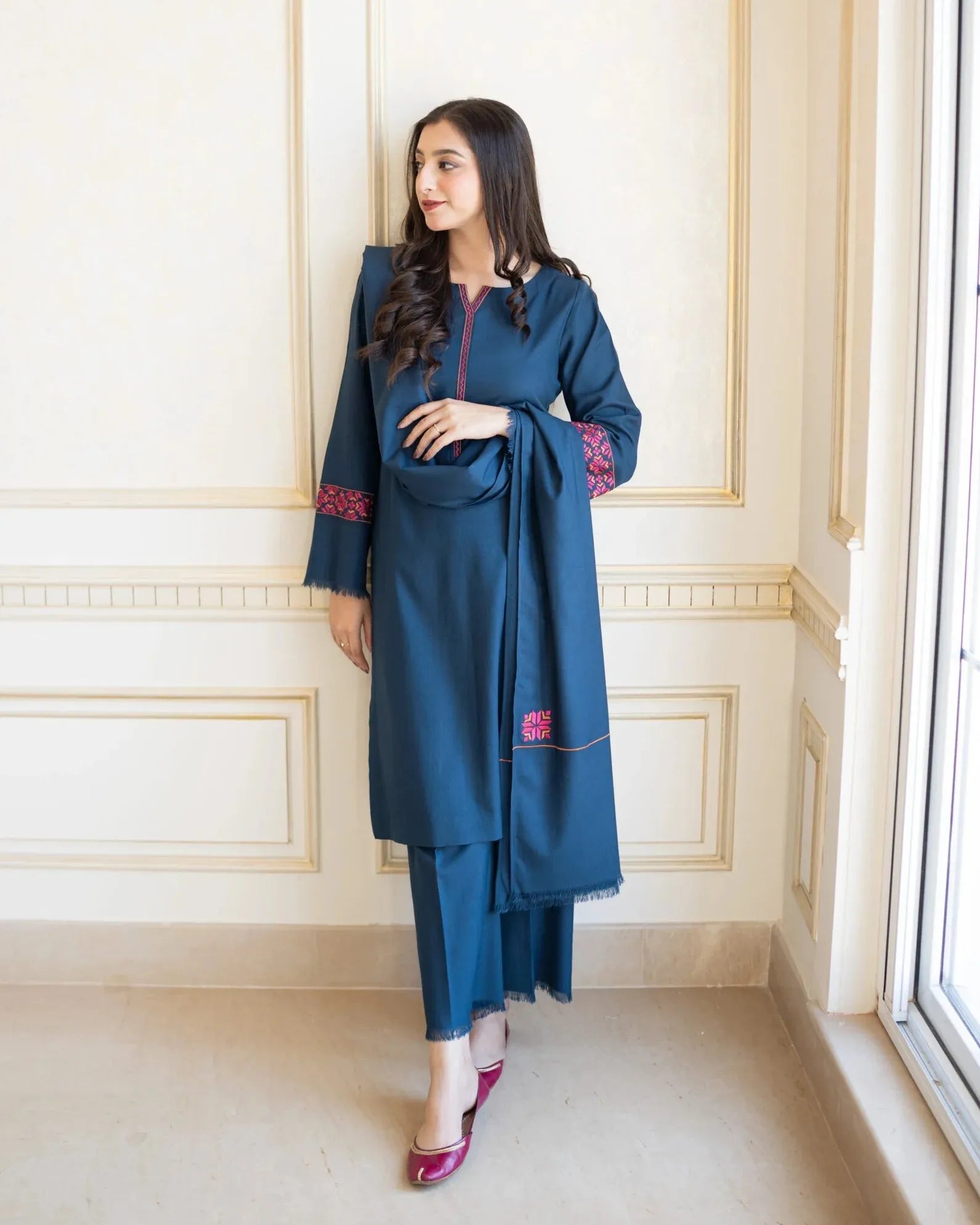 Woman wearing a blue traditional outfit with red embroidery in a room with white walls and a window.