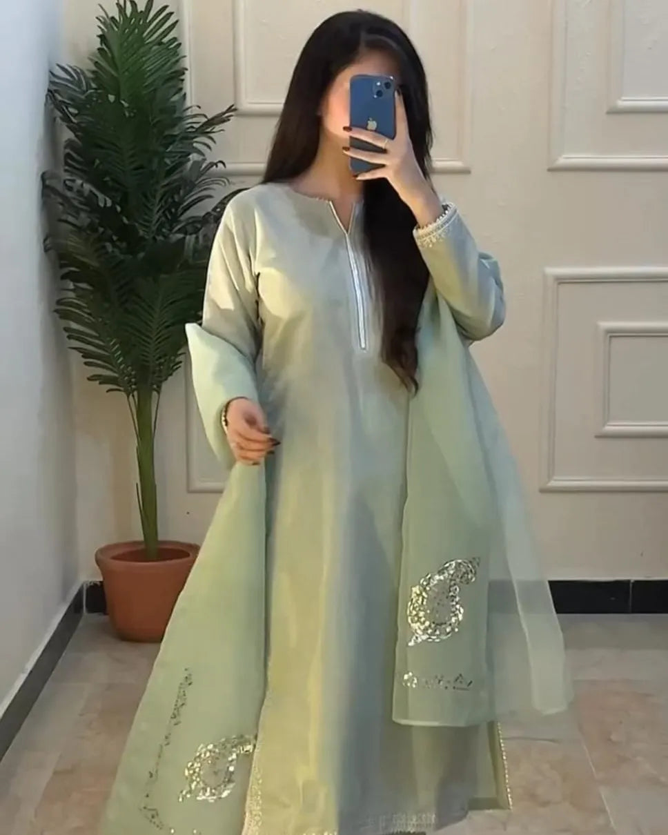 Person wearing a light green traditional outfit with embroidery, standing in a room with a plant and white paneled wall.