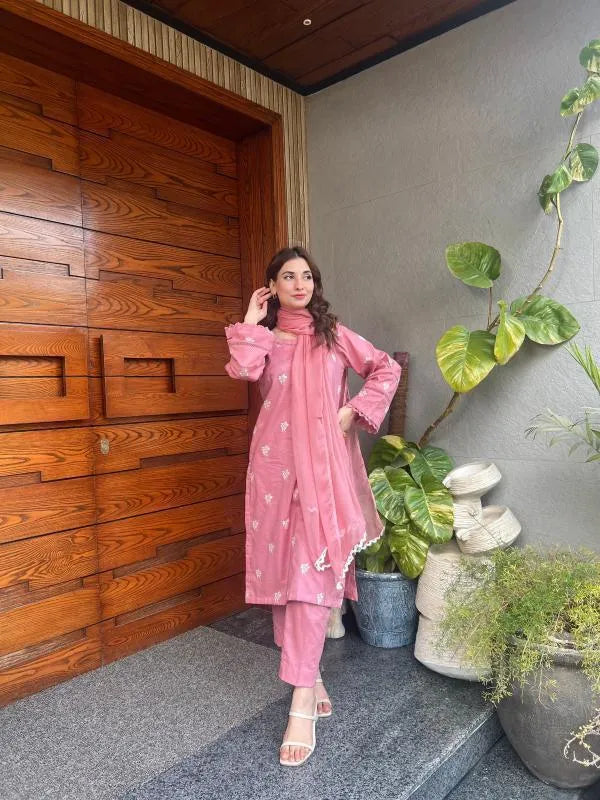 Woman in a pink traditional outfit standing in front of a wooden wall with plants.
