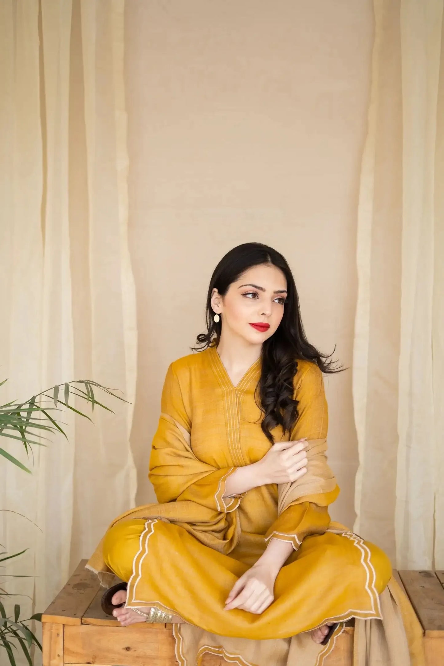 Woman in a mustard yellow traditional outfit sitting on a wooden bench against a beige curtain.