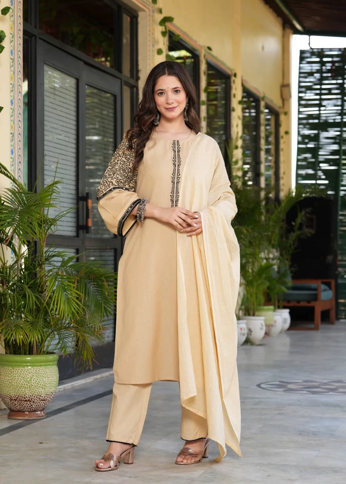 Woman in a beige traditional outfit standing in an outdoor setting with plants and a building in the background.
