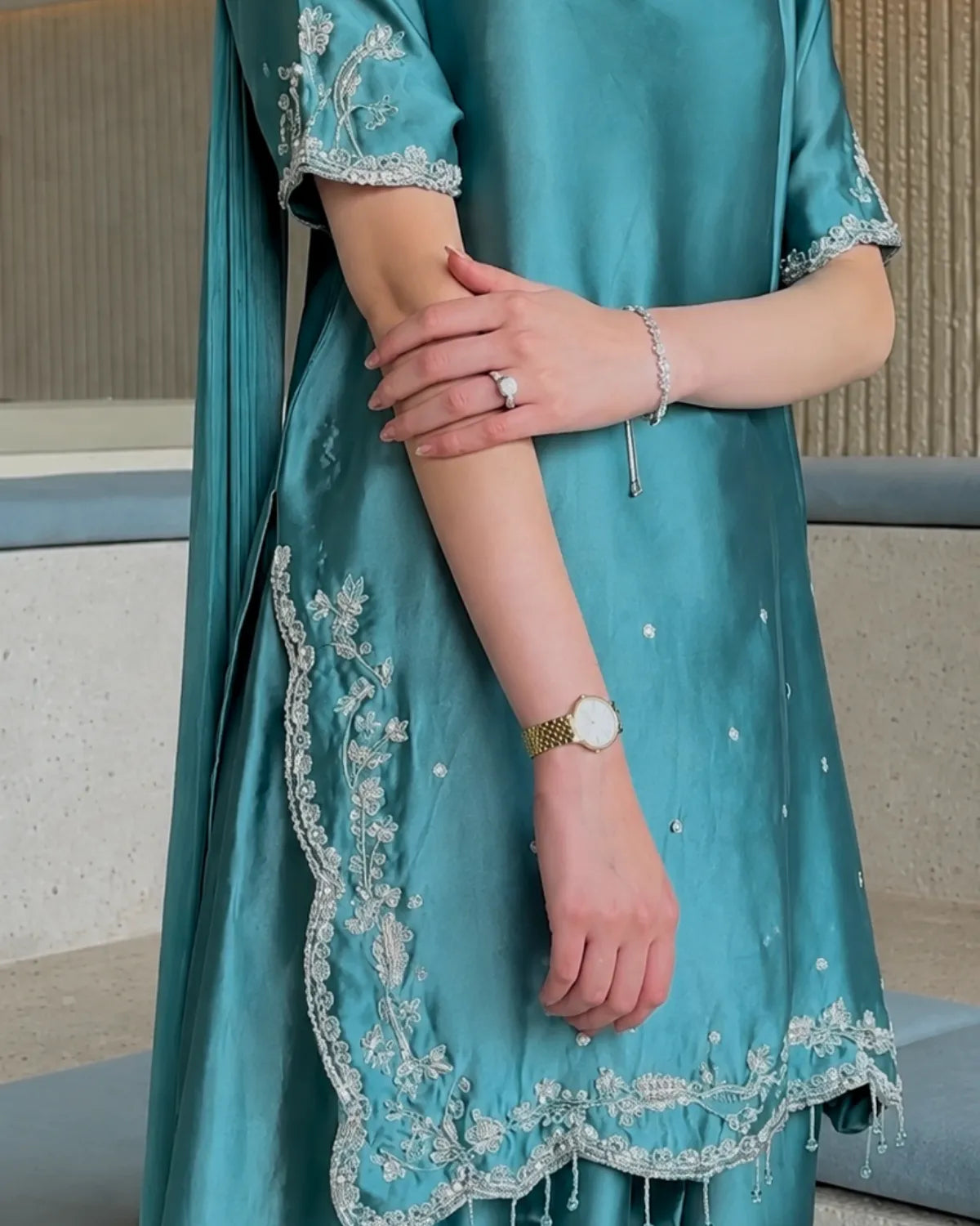 A close-up of a person wearing a teal blue satin kurta with intricate silver and white floral embroidery along the borders and sleeves, paired with a matching dupatta.