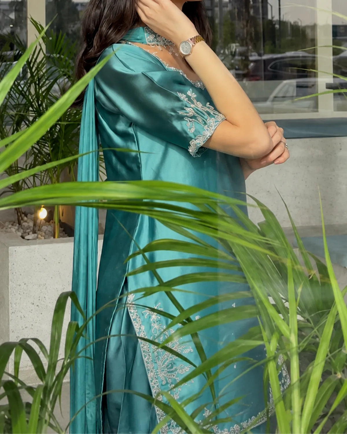 A woman wearing a teal satin kurta with silver floral embroidery, partially framed by green palm leaves in the foreground.