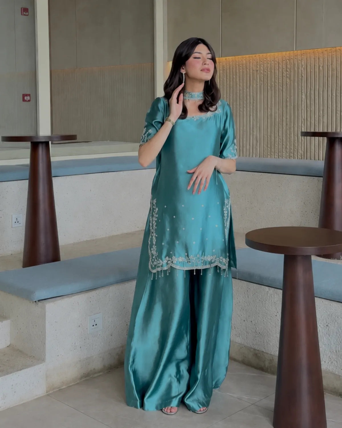 A woman models a teal green satin kurta and trouser set with silver embroidery, standing in a modern indoor setting with concrete steps and wooden tables.