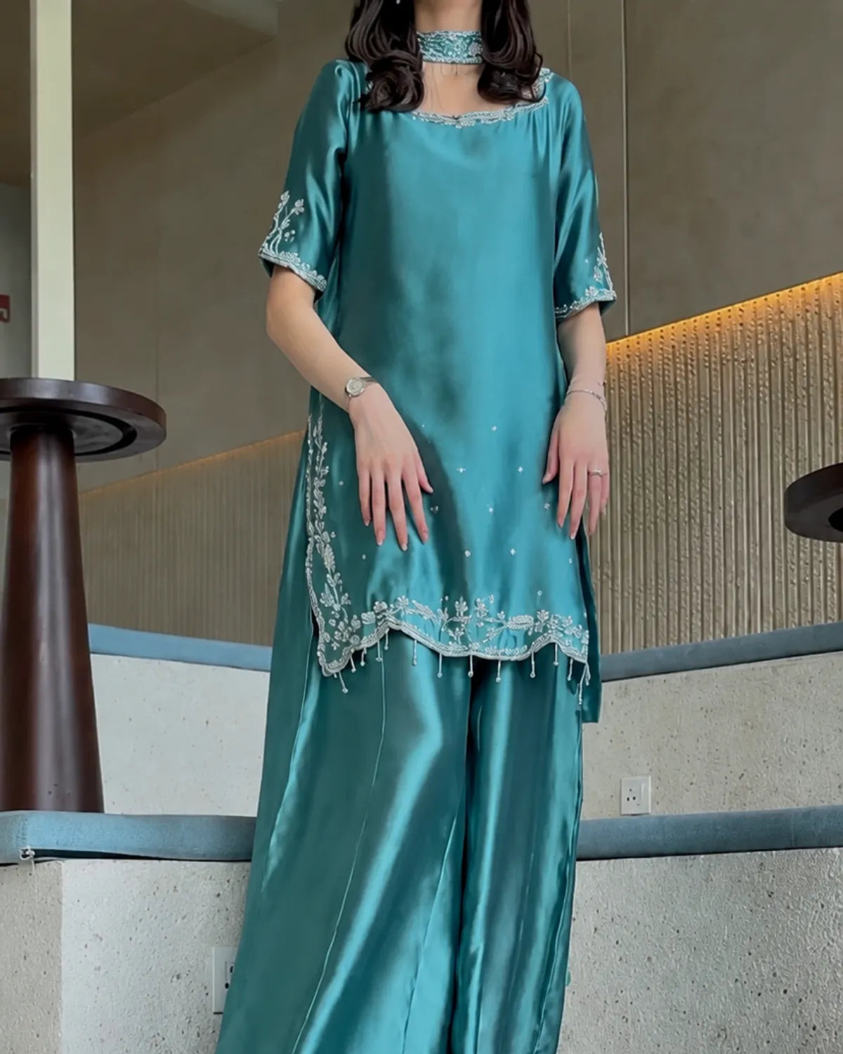 A woman wearing a teal satin kurta and wide-leg pants set with silver embroidery, standing on an indoor staircase.