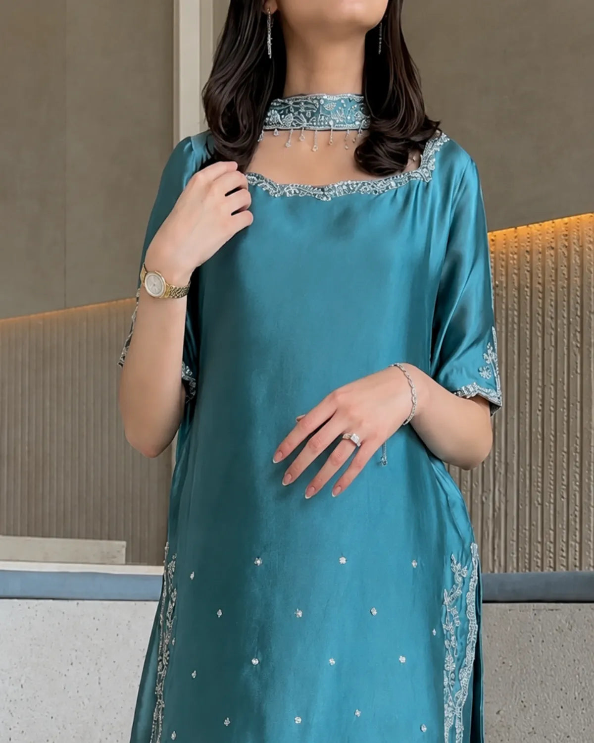A woman wearing a teal blue satin-like kurta with silver embroidery, a matching embroidered choker, and silver embellished sleeve cuffs, shown in a modern indoor setting.