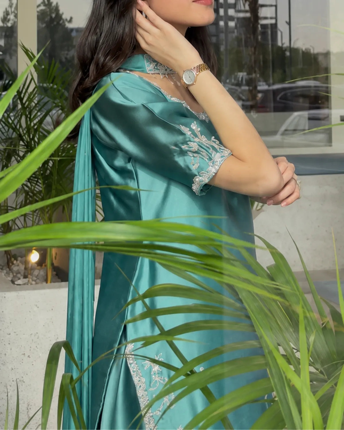 A woman wearing a teal satin kurta with silver floral embroidery and a matching dupatta, photographed in an outdoor setting with green palm leaves in the foreground.