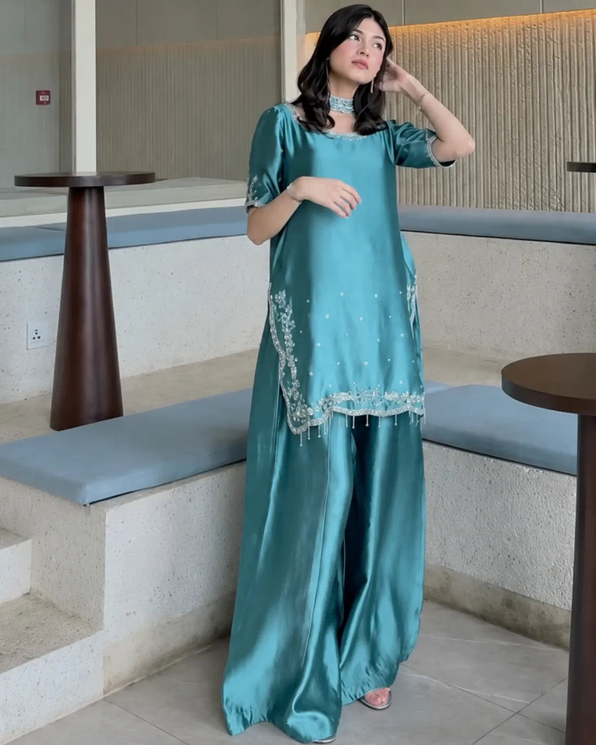A woman models a teal green satin kurta set with matching wide-leg trousers in a modern indoor setting with stone steps and wooden tables.