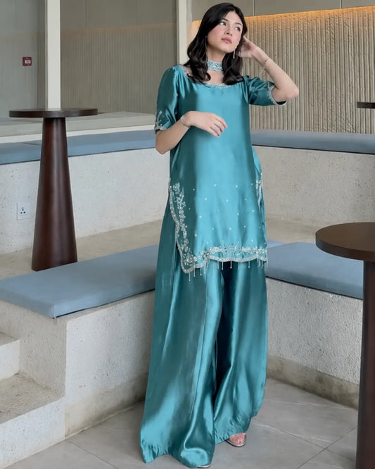 A woman models a teal green satin kurta set with matching wide-leg trousers in a modern indoor setting with stone steps and wooden tables.