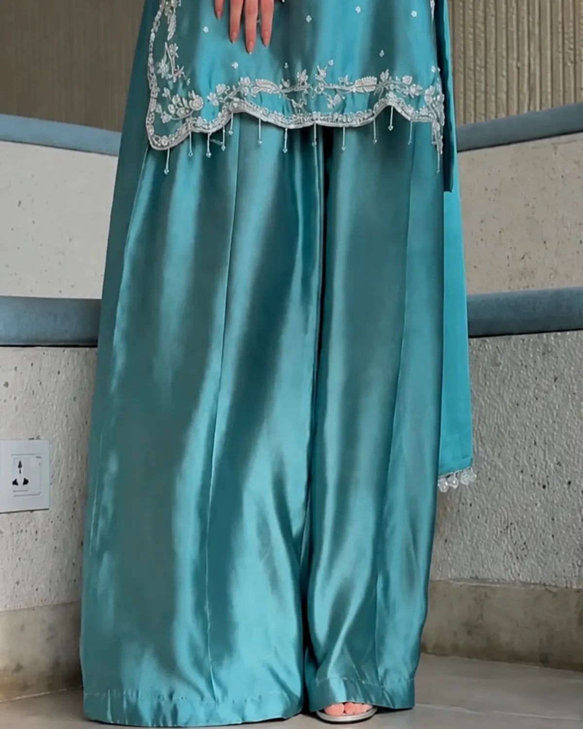 A close-up of a person wearing a teal satin three-piece outfit with wide-leg pants, a matching top, and silver embroidery along the hem.