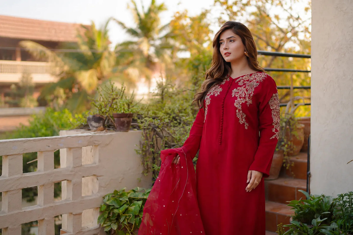 A woman wearing a long red embroidered kurta with a matching sheer red dupatta, standing on an outdoor balcony with plants and stairs in the background.