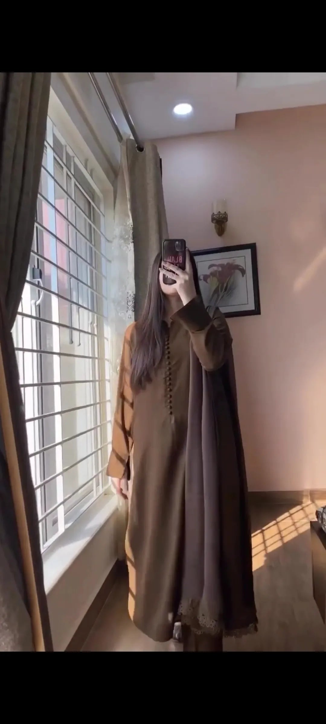 Person taking a mirror selfie wearing a long, light-colored dress in a room with a window and framed picture on the wall.