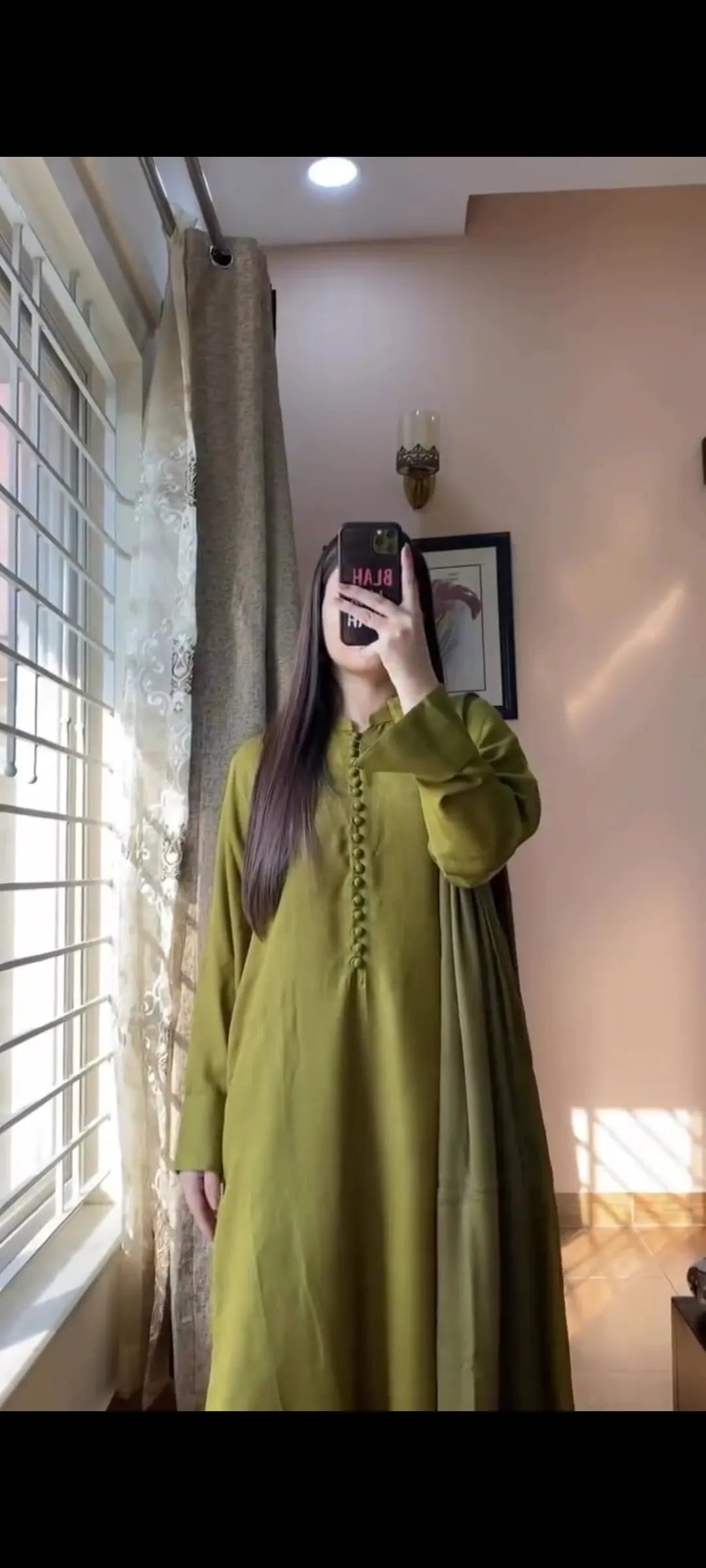Person wearing a green dress taking a mirror selfie in a room with a window and curtain.