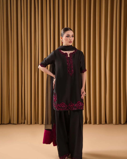 Woman wearing a black outfit with red embroidery in front of a beige curtain