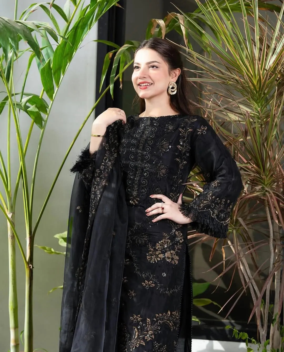 Woman wearing a black embroidered dress with a sheer overlay in front of plants.