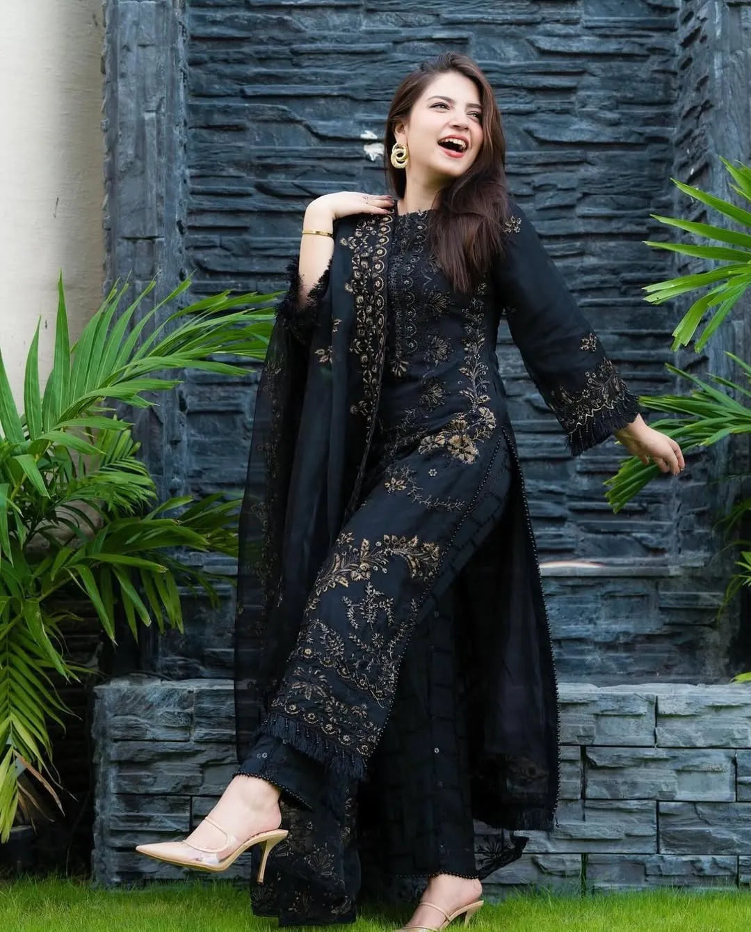 Woman in a black embroidered outfit standing against a stone wall with greenery.