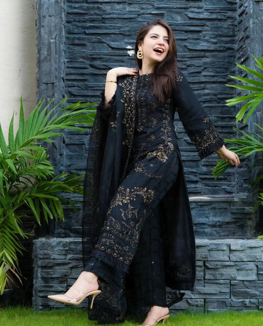 Woman in a black embroidered outfit standing against a stone wall with greenery.