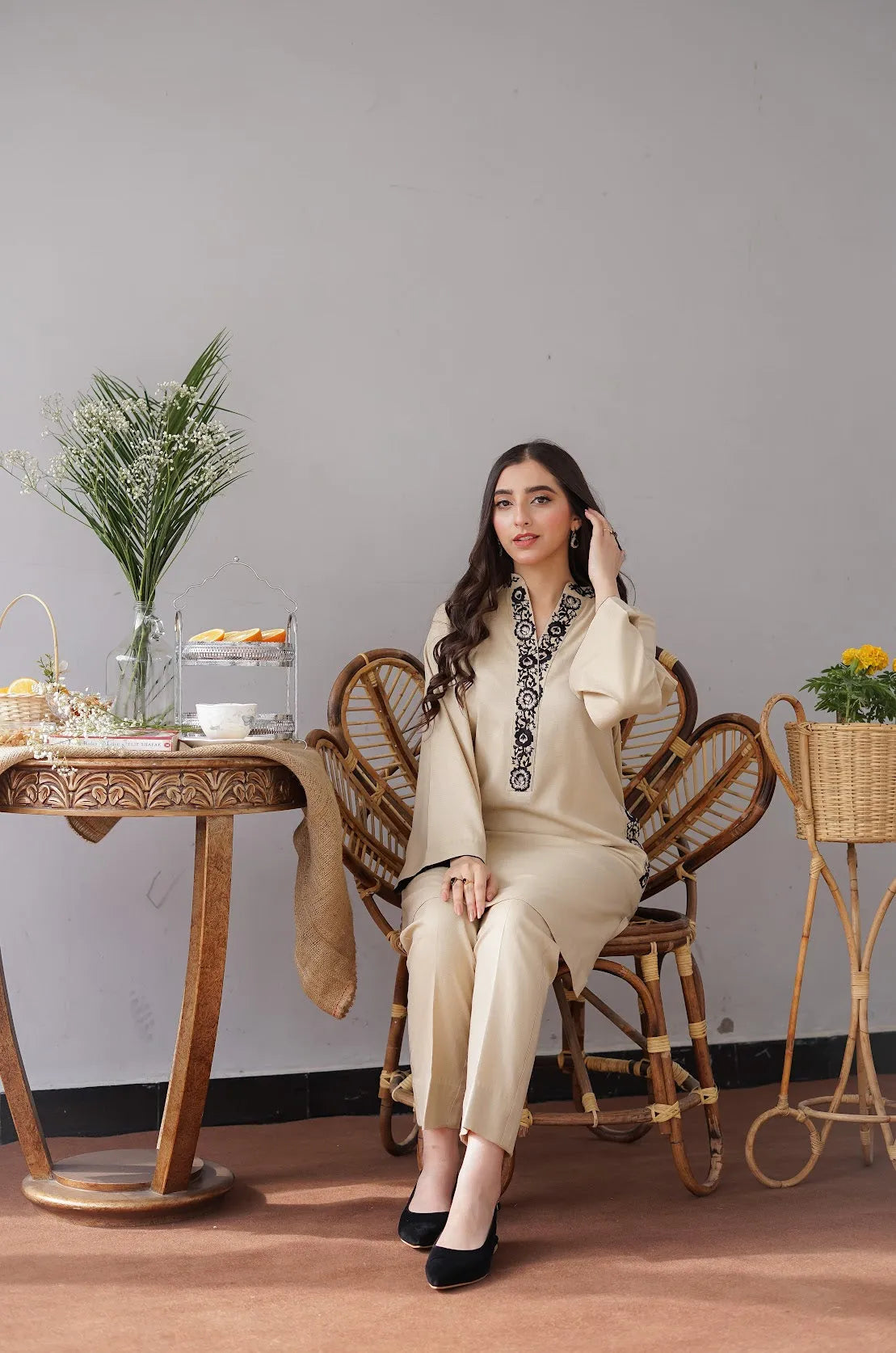 Woman sitting on a wicker chair in a room with a wooden table and decorative items.