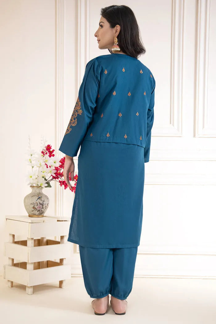 Woman wearing a blue traditional outfit with floral embroidery, standing in a minimalistic room.