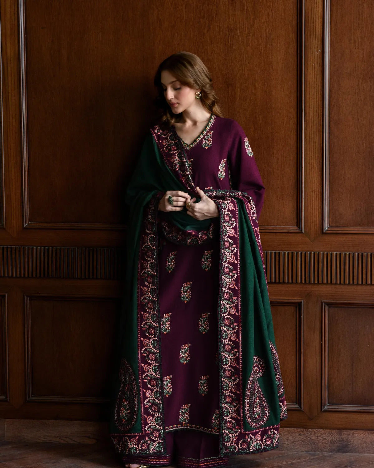 Woman wearing a long, dark purple and green embroidered dress against a wooden paneled wall.