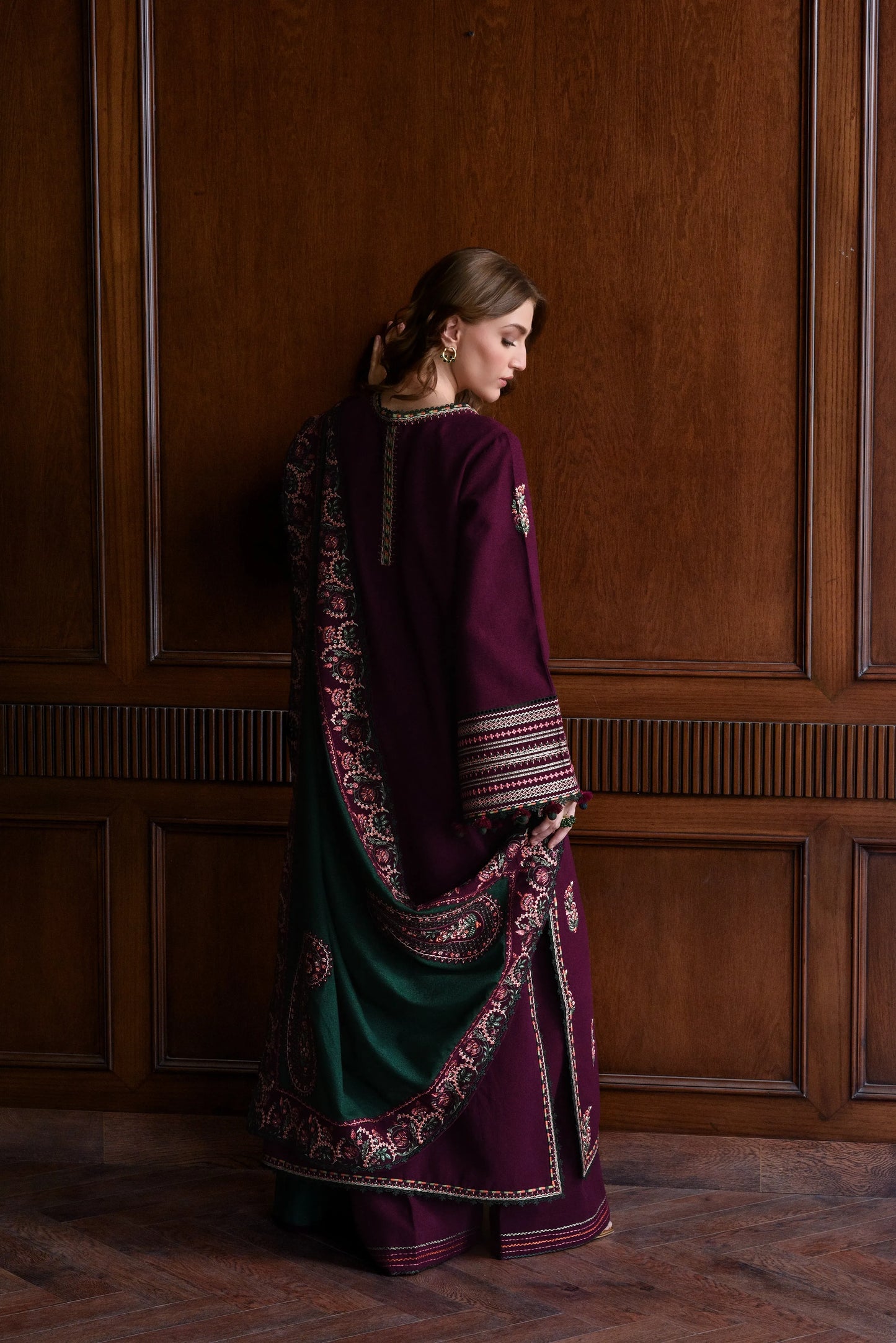 Woman wearing a traditional outfit with a richly patterned shawl against a wooden paneled wall.