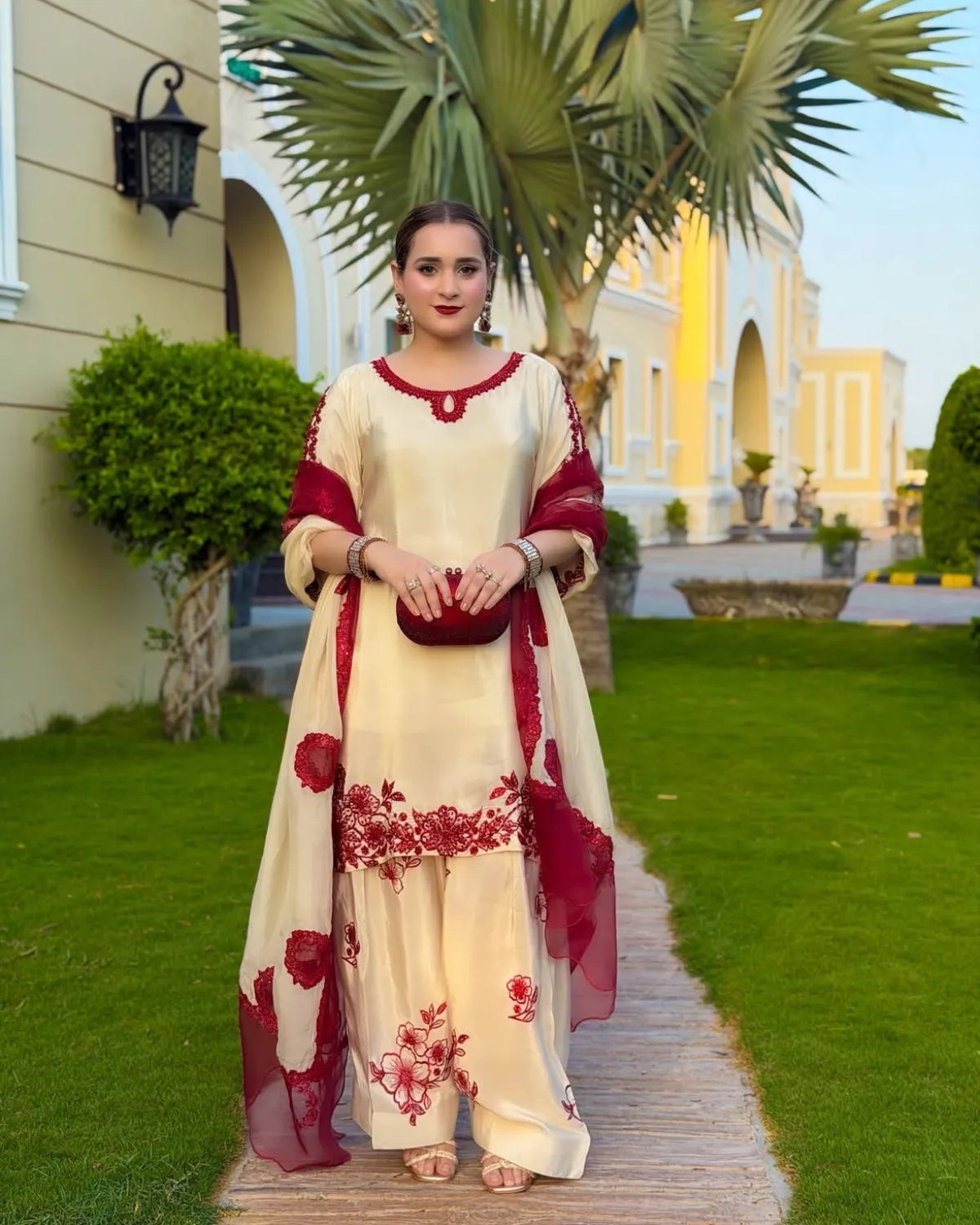 A woman stands on a wooden path in a garden setting, wearing a cream-colored traditional outfit with deep red floral embroidery and matching red dupatta.