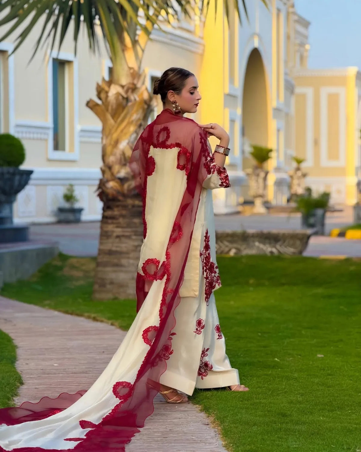 A woman stands outdoors on a paved path beside a green lawn, wearing a cream-colored traditional outfit with deep red floral embroidery and a matching sheer red dupatta. The background features a large yellow building, palm trees, and manicured grass.