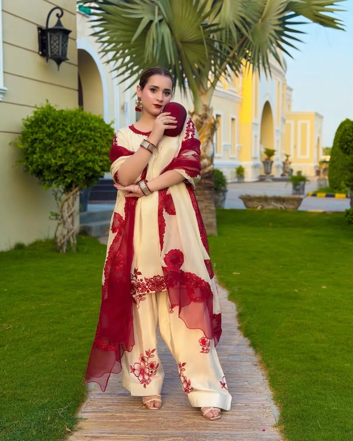 A woman stands on a wooden path in a landscaped garden wearing a cream and deep red embroidered traditional outfit with a matching sheer dupatta.