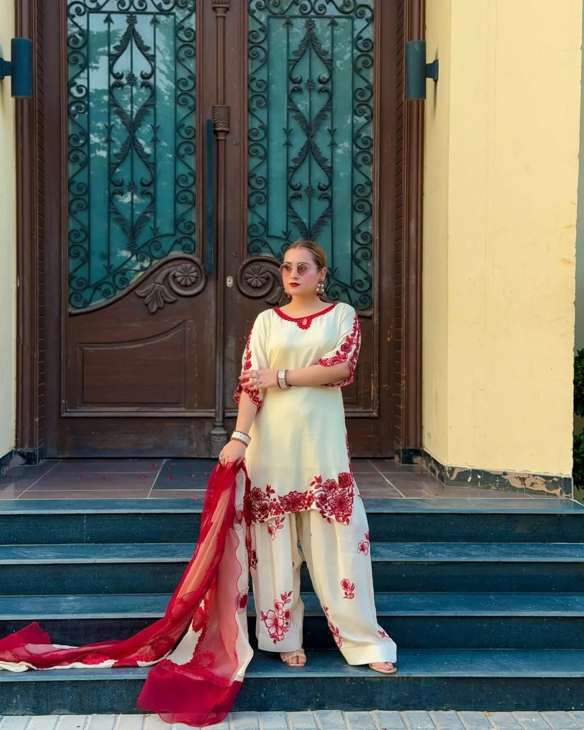 A woman stands on outdoor stone steps in front of a large ornate brown door, wearing a cream-colored embroidered kurta set with red floral patterns and holding a matching red dupatta.