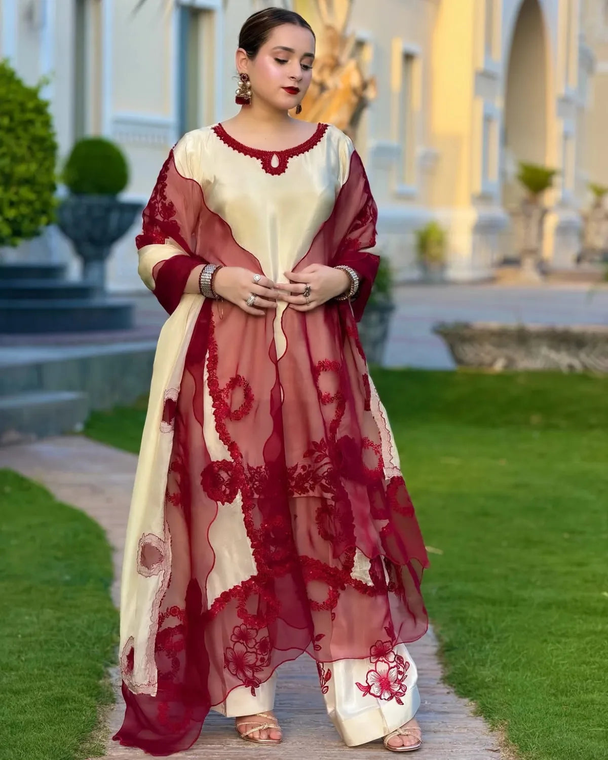 A woman stands outdoors on a garden path wearing a traditional outfit with a cream and deep red color-blocked design, sheer red embroidered overlays, and matching red pants.
