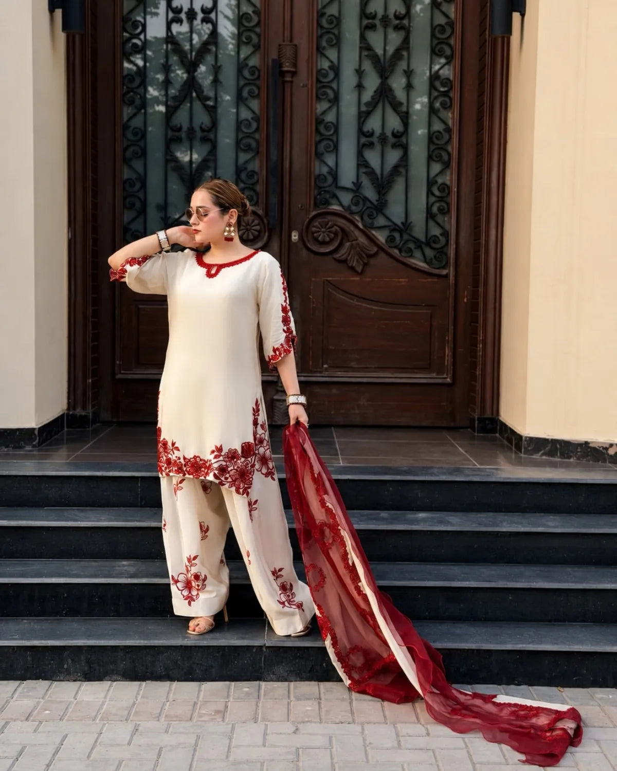 A woman stands on black stone steps in front of a dark wooden door, wearing a cream-colored traditional outfit with red floral embroidery and holding a matching red dupatta.