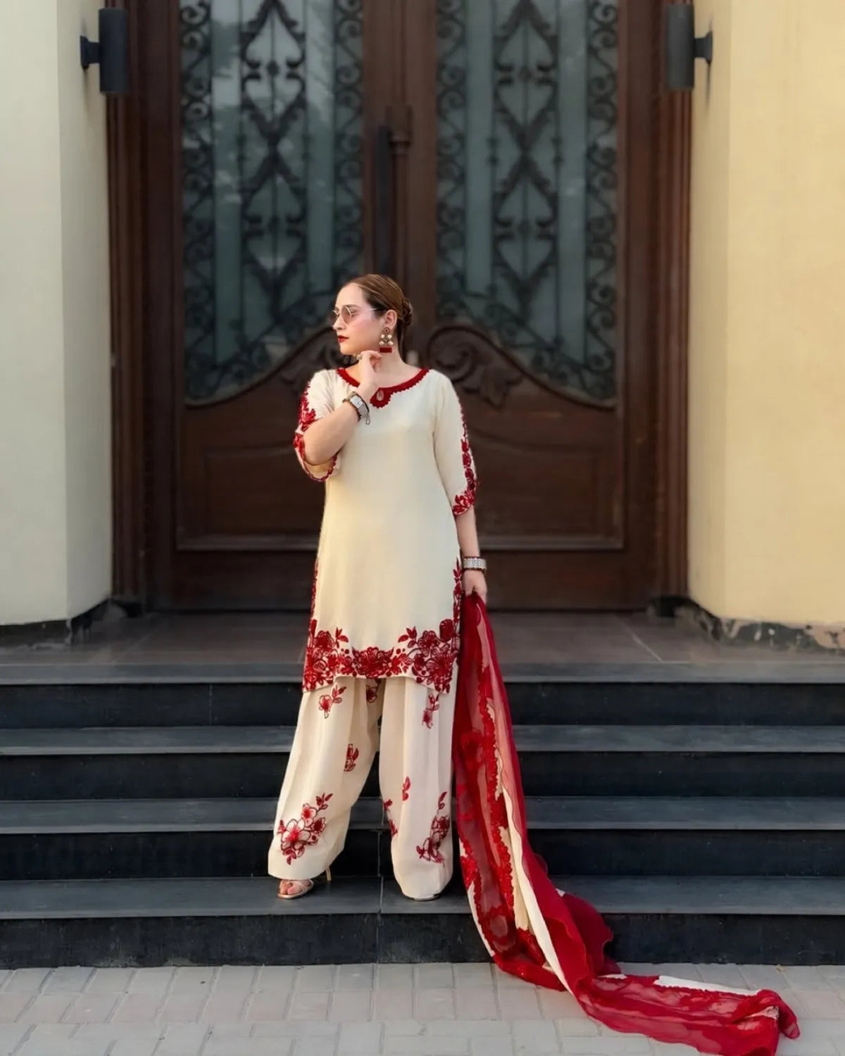 A woman models a cream-colored embroidered kurta and trouser set with a red dupatta, standing on steps in front of a dark wooden door.