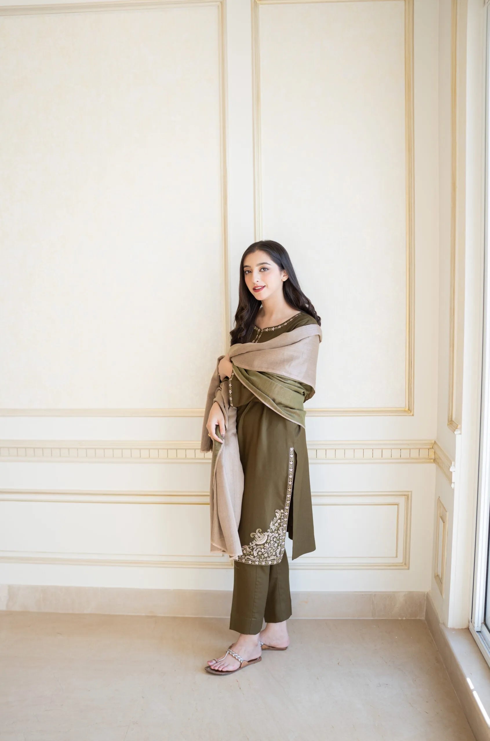 Woman in a green outfit with a beige shawl standing against a white wall.