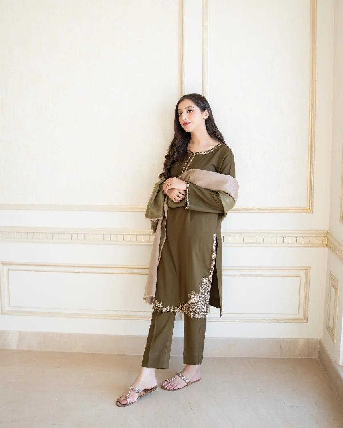 Woman in an olive green traditional outfit standing against a white wall.