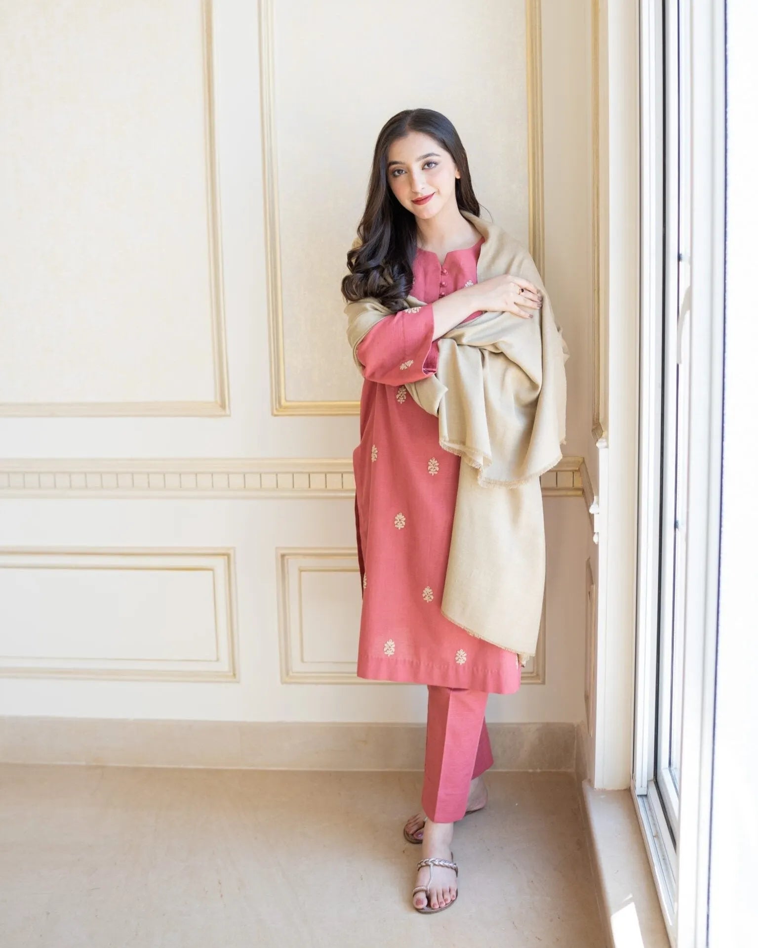 Woman in a pink traditional outfit with a beige shawl standing in a room with white walls and a window.