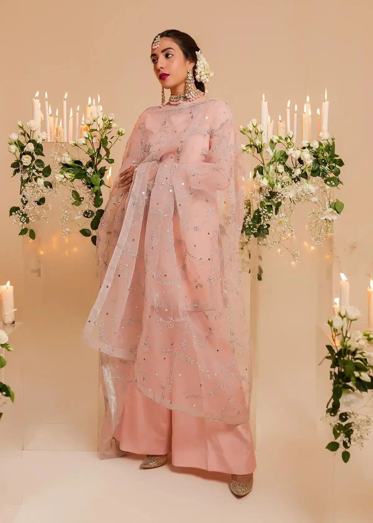 Woman in a pink traditional outfit standing in a decorated room with candles and flowers.